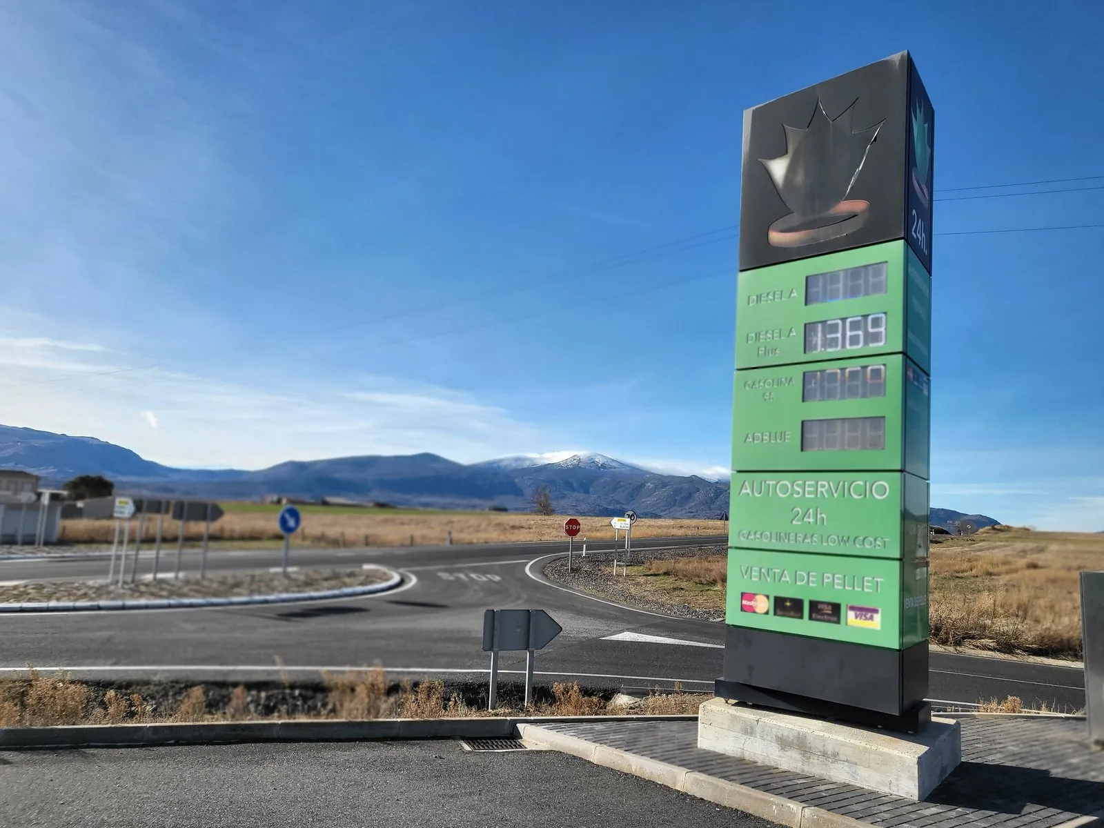 Cartel de precios de una gasolinera low cost en La Torre (Ávila): el panel verde marca 1,369 en una de las líneas de gasóleo, con la sierra nevada al fondo