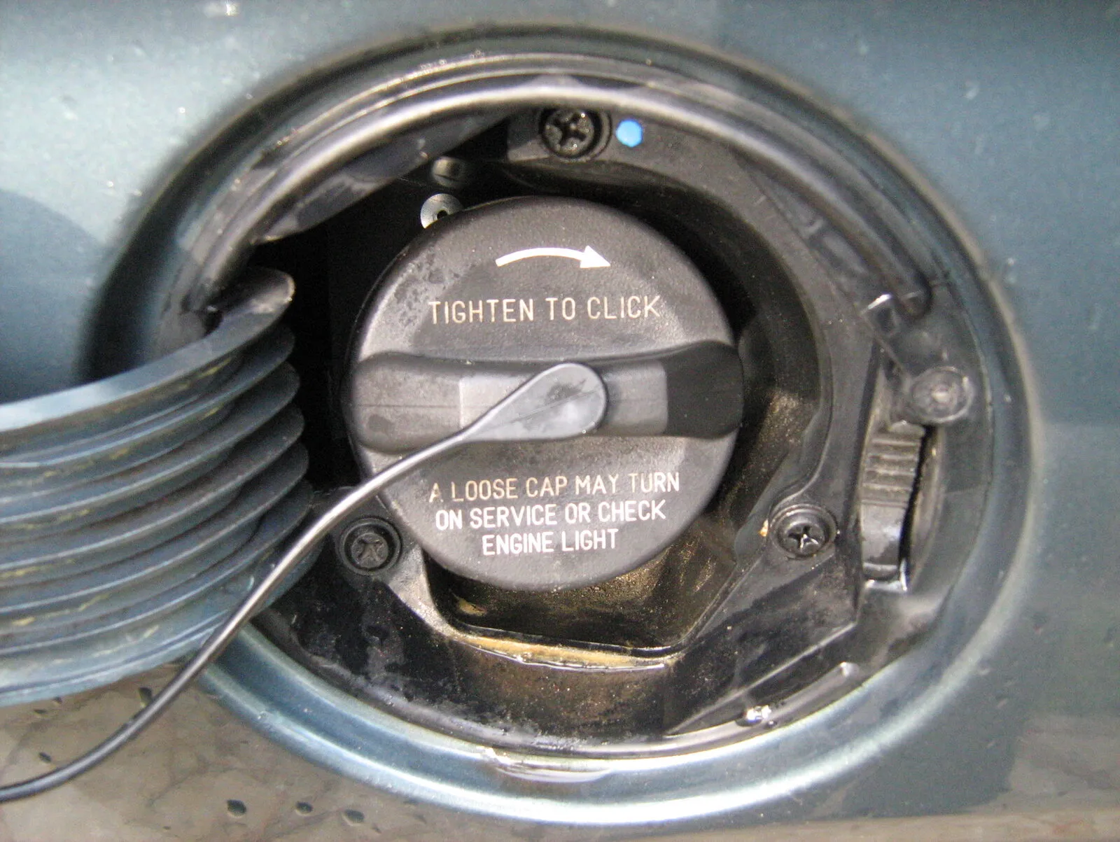 An open fuel filler door with a black gas cap printed “Tighten to click — a loose cap may turn on service or check engine light”